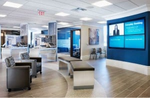 Image of a Bank Branch. Wood floors, desks, and chairs along a wall. Digital signage hanging on wall.